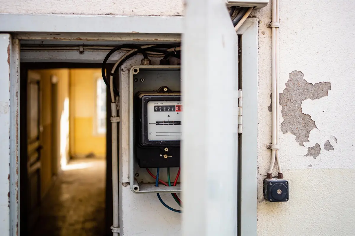 Compteur électrique avec câbles visibles dans un boîtier mural, couloir en arrière-plan, peinture écaillée.
