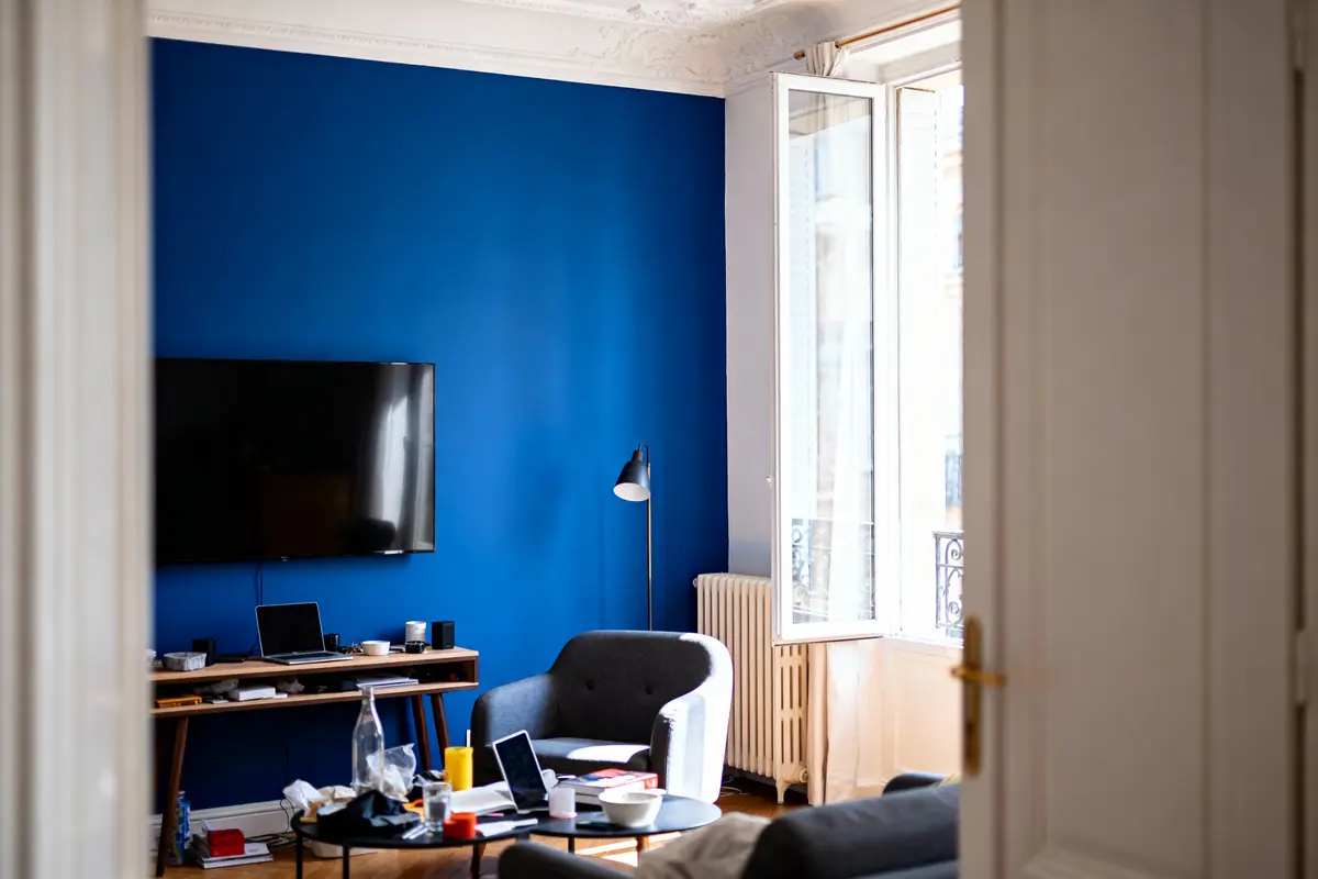 Salon moderne avec mur bleu, canapé gris, TV, bureau encombré, grande fenêtre ouverte.