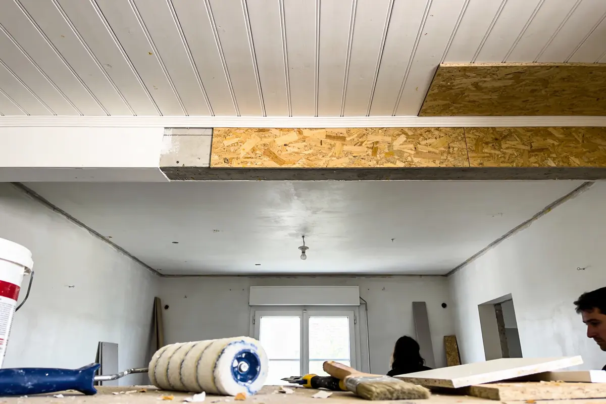 Salle en rénovation avec plafond en lambris, outils de peinture et bricolage visibles sur le plan de travail.