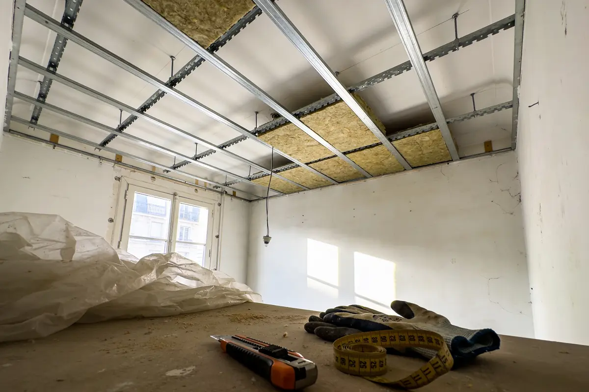 Construction intérieure avec plafond métallique, laine isolante, fenêtre, couteau, gants, mètre ruban sur table en bois.