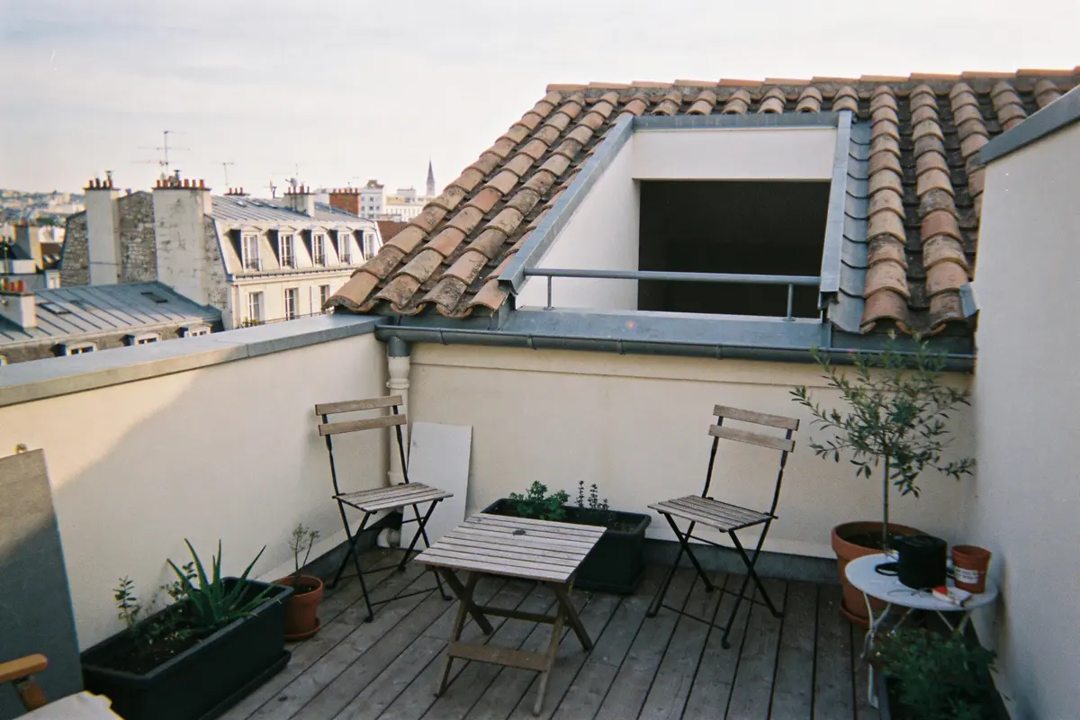 Terrasse parisienne sur un toit avec tables, chaises pliantes, plantes en pots, vue sur bâtiments voisins.