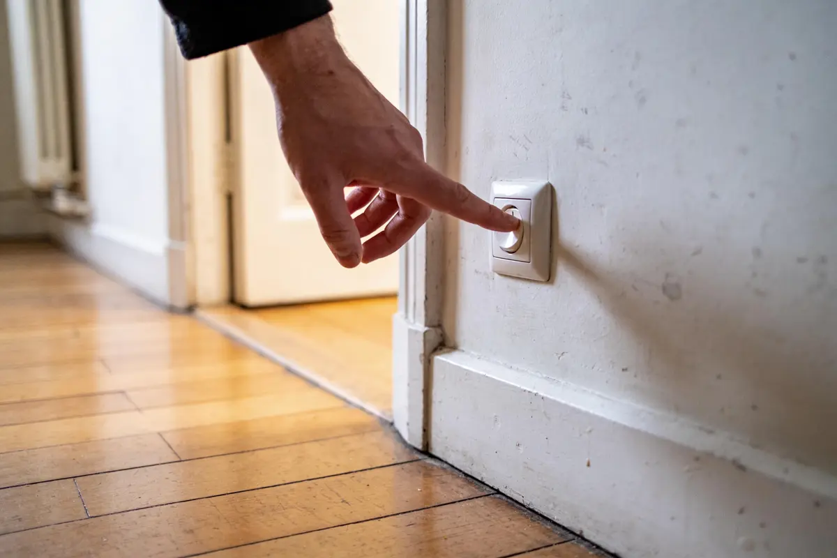 Une main touche un interrupteur lumineux sur un mur blanc, parquet en bois en arrière-plan.