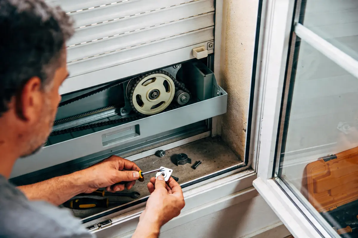 Un homme répare un volet roulant près d'une fenêtre ouverte avec des outils.