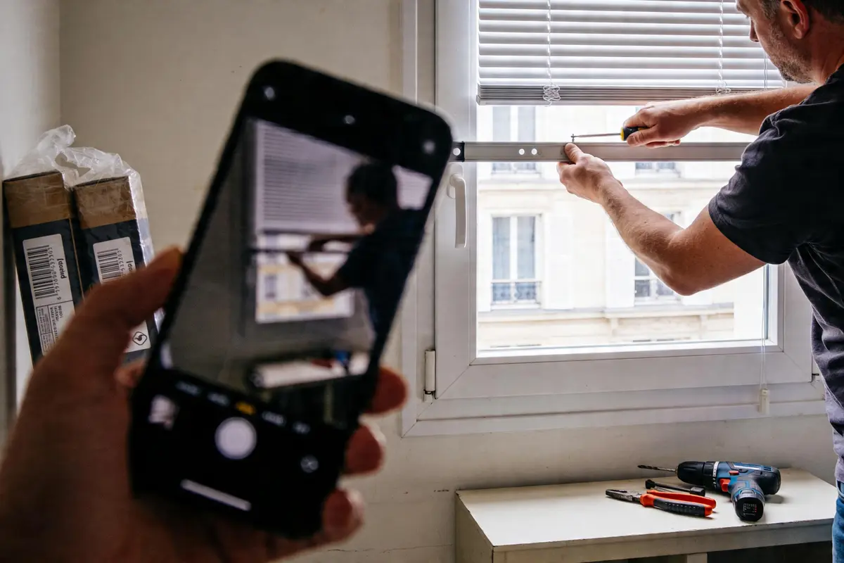 Homme installe un store devant une fenêtre, capturé via un smartphone, outils posés sur une table.
